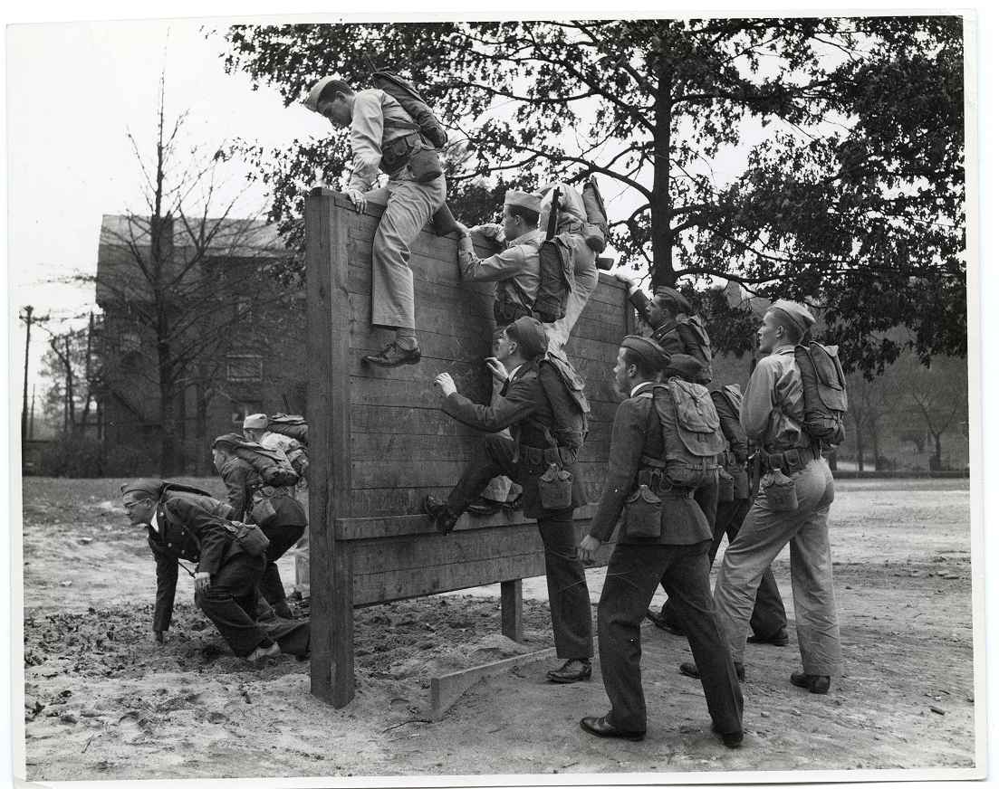 A group of soldiers in uniform practice teamwork by climbing over a wooden obstacle in an outdoor training area, conveying determination and cooperation.