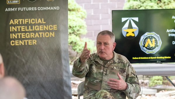 A U.S. Army general in uniform gestures while speaking at the Army Futures Command's Artificial Intelligence Integration Center. A presentation screen displays logos in the background, conveying a focus on technology and innovation. The setting is outdoors with a relaxed, informative atmosphere.