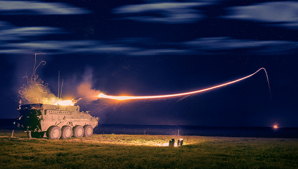 An armored military vehicle at night fires a bright missile, creating a vivid arc of light against a starry sky, evoking a sense of power and precision.