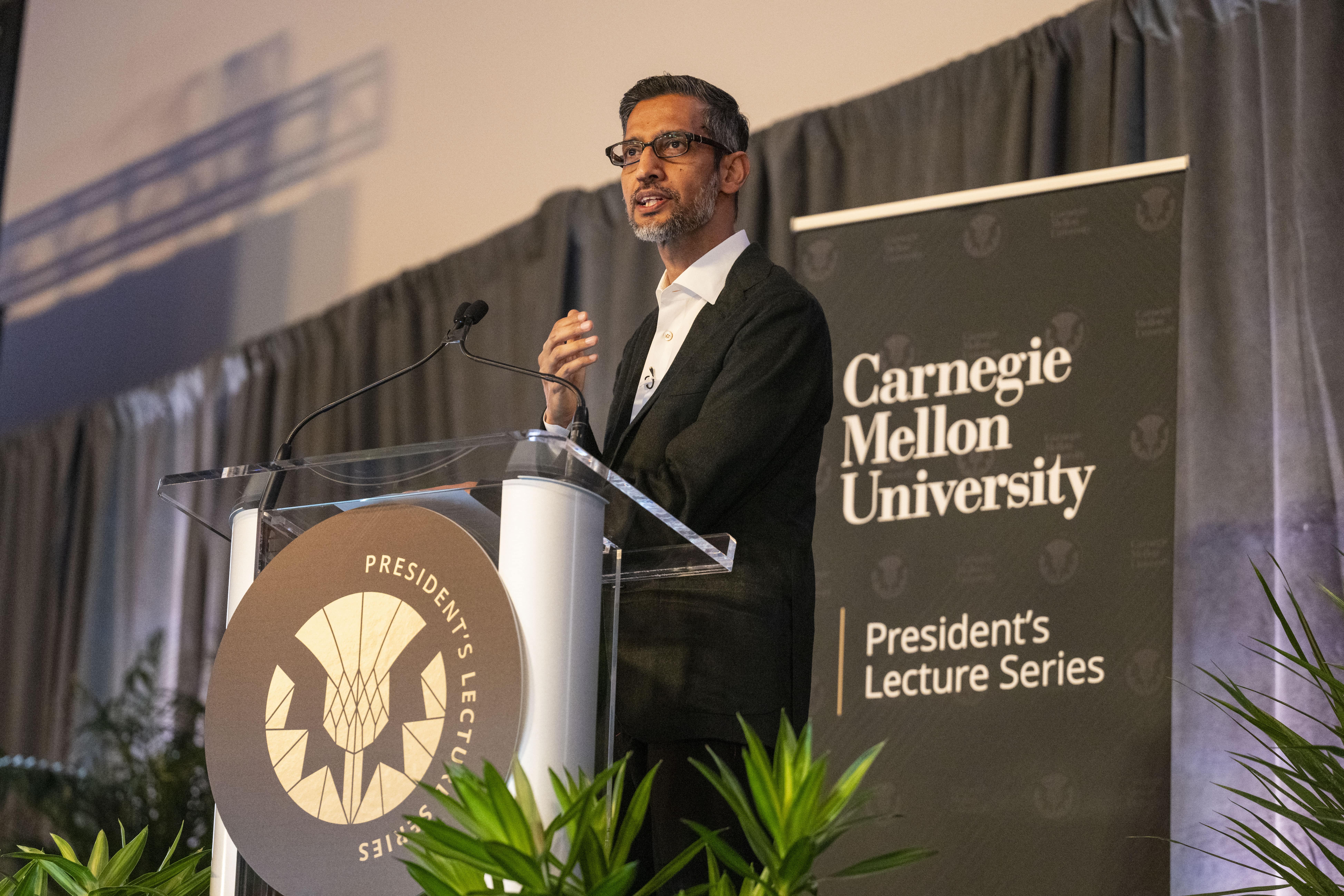 Pichai stands at a clear acrylic podium with a black Carnegie Mellon University President's Lecture Series sign behind him.