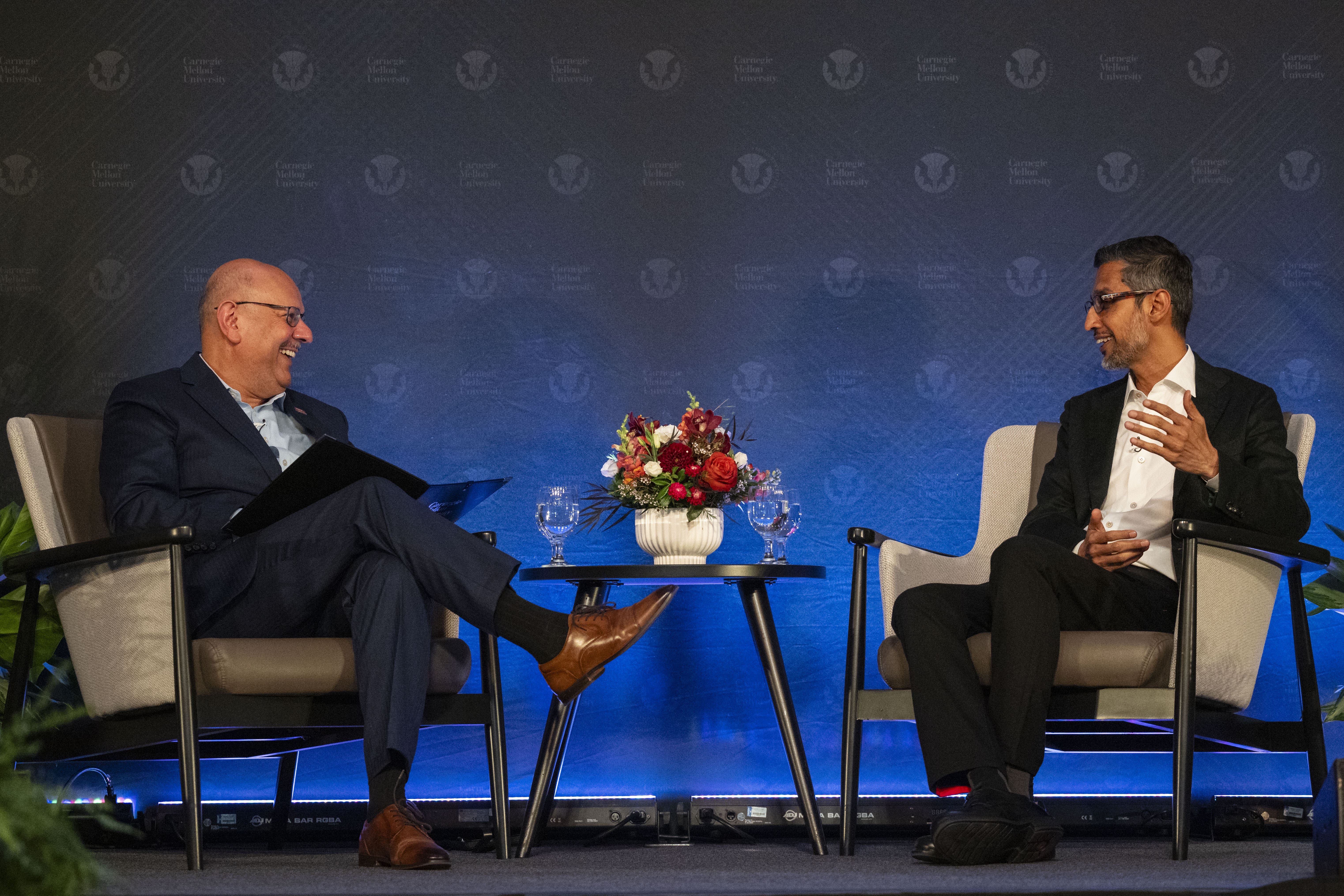 Jahanian (left) and Pichai (right) sit on beige chairs in front of a dark blue background..
