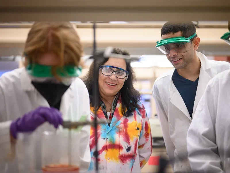 In a lab, a professor wearing a tie dye coat interacts with students in white coats. They are all wearing protective glasses.