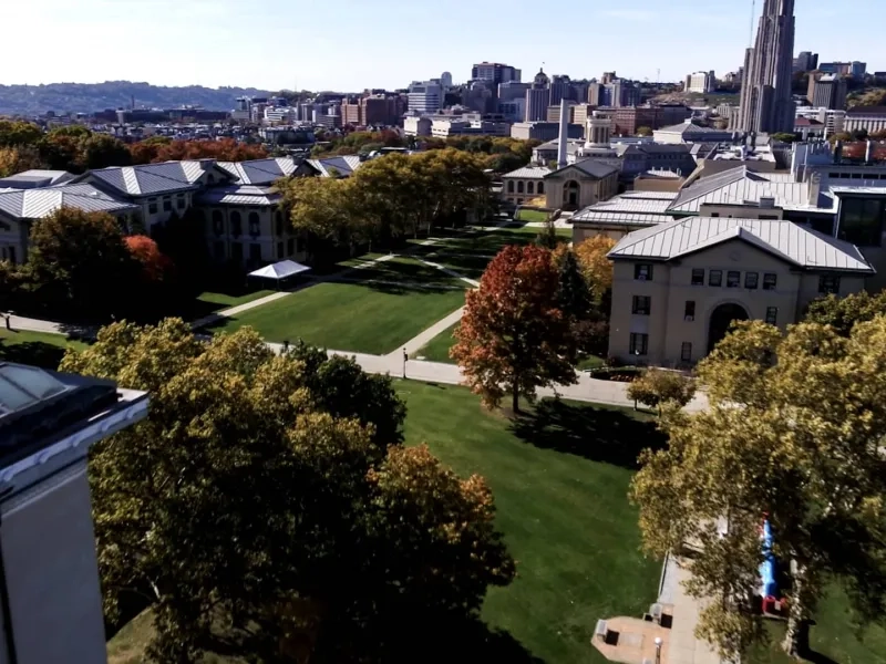 Aerial view of CMU's campus with green lawns and tree-lined paths and the Pittsburgh cityscape in the background