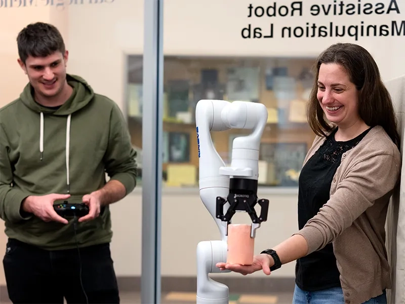 Student controlling a robotic arm while their professor holds out their hand holding a ceramic cylinder.