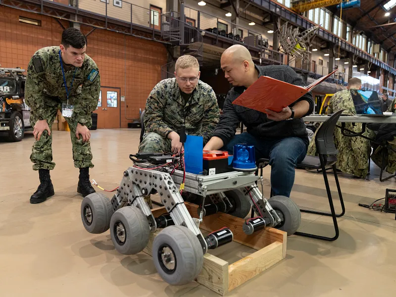 Three people in military attire and a civilian examine a small wheeled robot in an industrial setting. They appear focused and engaged in discussion.
