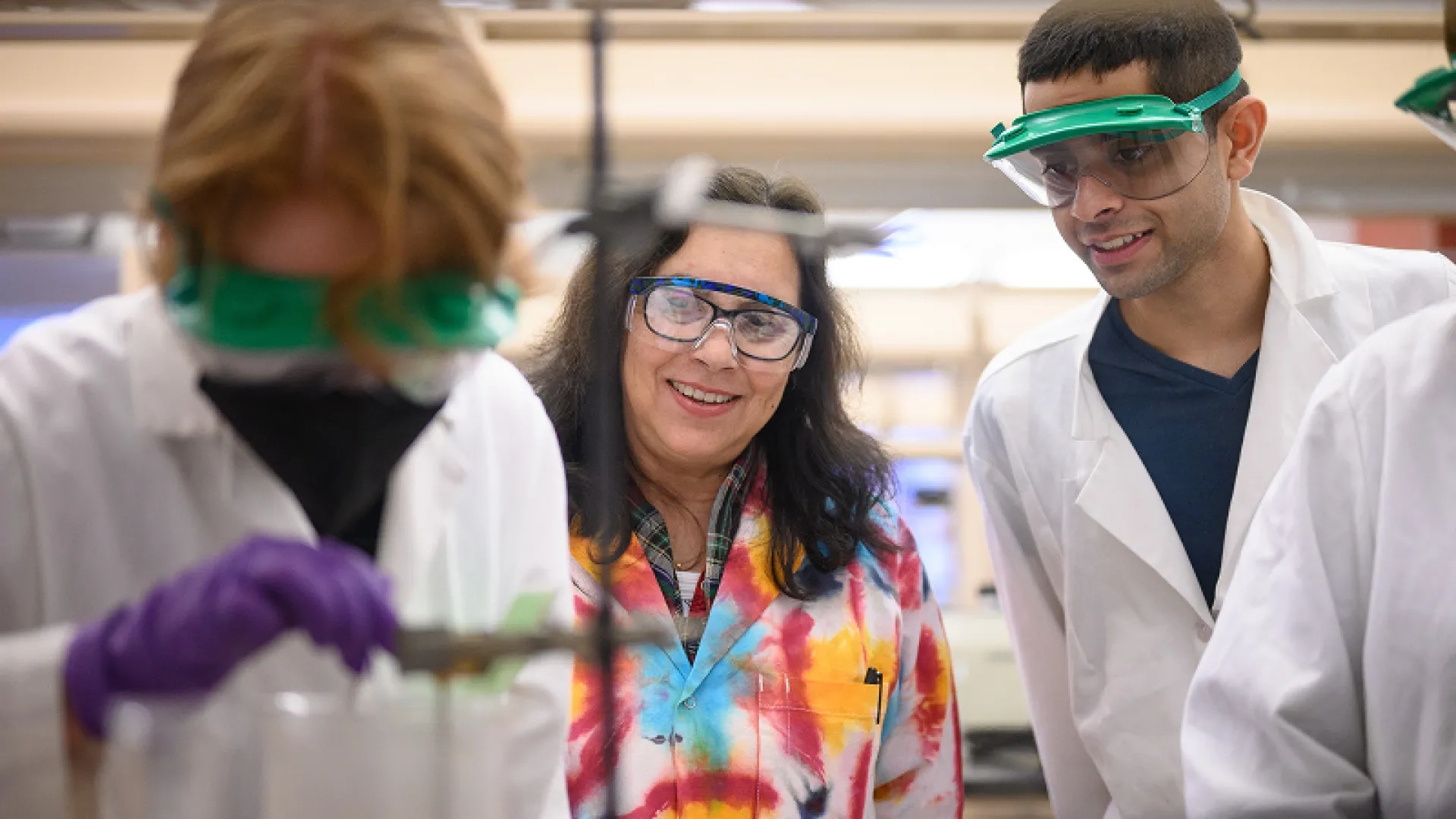 In a lab, a professor wearing a tie dye coat interacts with students in white coats. They are all wearing protective glasses.