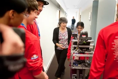 Several people watching as a young girl pushes a cart holding machines