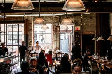 A bustling industrial-style café with exposed brick walls and large windows as patrons sit at tables enjoying drinks and meals