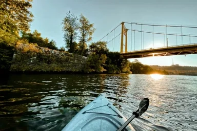 View from a white kayak on a river outside of Pittsburgh at sunset with a suspension bridge ahead and greenery on the riverbanks