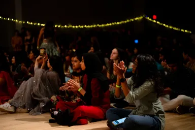 Several people sitting on the floor clapping with a string of lights overhead